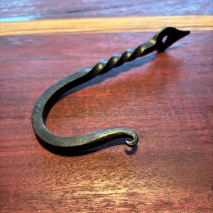 May include: A black metal hook with a twisted stem and a decorative loop at the end. The hook is handcrafted and has a rustic finish.