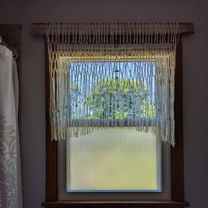 May include: A white macrame curtain hangs in a window with a view of a green tree and blue sky. The window has a wood frame and a frosted glass pane.