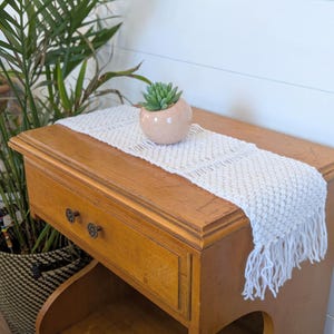 May include: A white crocheted table runner with fringe on a wooden table with a drawer. A small pink planter with a succulent plant is on the table runner.