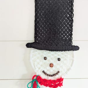 May include: A white yarn snowman wall hanging with a black yarn hat, brown bead eyes and nose, and a red and green yarn scarf.