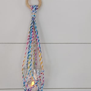 May include: A colorful macrame candle holder with a wooden ring at the top. A small white candle is burning inside a glass jar.
