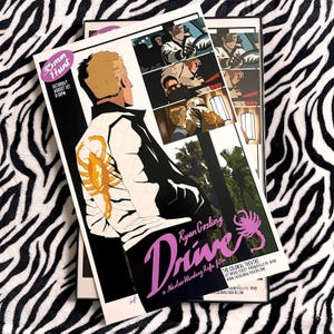 May include: A rectangular movie poster for Drive featuring a blonde character wearing a white jacket with a gold scorpion emblem on the back. The poster includes comic-style panels, the text Drive, Ryan Gosling, and The Colonial Theatre details.