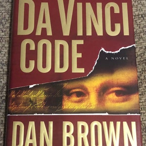 Op de afbeelding: Een rode boekomslag met gouden letters die "The Da Vinci Code" en "A Novel" lezen. De naam van de auteur, "Dan Brown", staat in gouden letters onder de titel. De boekomslag toont ook een close-up van een schilderij met de ogen van een vrouw, en de tekst "Auteur van Angels & Demons" is in gouden letters onderaan gedrukt.