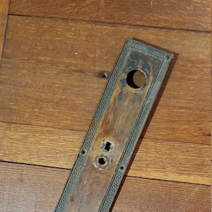 May include: An antique door plate with a rectangular shape and a decorative Greek key border. The plate has a weathered, aged appearance with holes for a doorknob and key. The metal has a dark, tarnished finish.