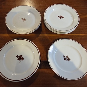 May include: A collection of white ceramic plates with a brown leaf design in the center. Each plate has a brown rim. The plates are arranged on a dark wood surface.