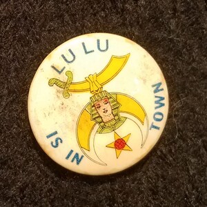 May include: A round, off-white pin with the text "LULU IS IN TOWN" in blue. The pin features a gold and yellow design with a sword, a crescent moon, and a star. A woman's face is in the center of the design.