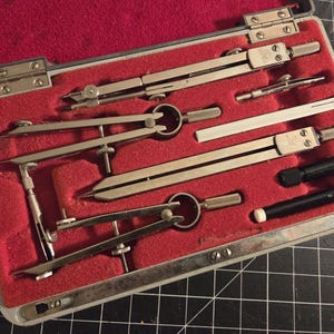 May include: A vintage drafting set in a metal case with a red velvet interior. The set includes several silver-colored compasses, dividers, and other drawing tools. The case is open, revealing the tools neatly arranged.