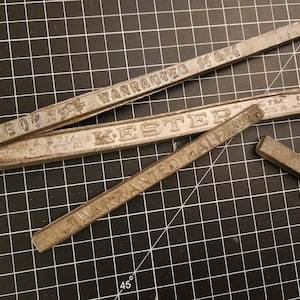 May include: Several rectangular, silver solder bars are arranged on a black grid. The bars have stamped text, including "WARRANTED" and "KESTER." One bar is broken. The image is taken from above.