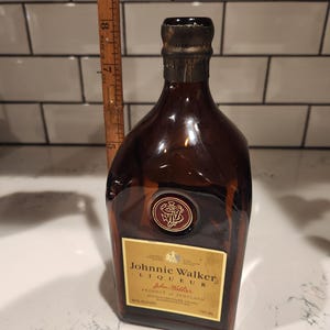 Pode incluir: Uma garrafa de vidro castanho de licor Johnnie Walker. A garrafa tem um r&oacute;tulo dourado com texto preto e um selo vermelho. A garrafa tem aproximadamente 23 cm de altura.