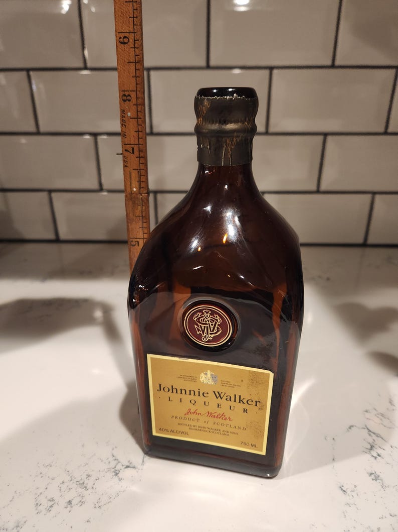 Pode incluir: Uma garrafa de vidro castanho de licor Johnnie Walker. A garrafa tem um r&oacute;tulo dourado com texto preto e um selo vermelho. A garrafa tem aproximadamente 23 cm de altura.