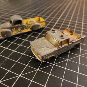 May include: Two vintage toy pickup trucks. The trucks are primarily gray with yellow and orange accents, showing signs of wear. One truck has a yellow bed, while the other has an open bed. The toys are set against a black grid background.