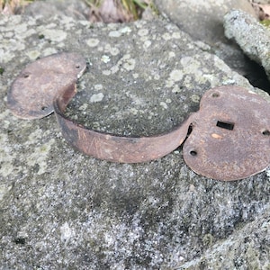 May include: An aged metal handle, likely for a cabinet or drawer, with two oval plates and a curved handle. The handle shows signs of rust and wear, indicating its age. The hardware is a dark brown colour.