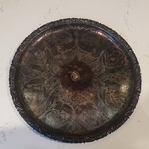 May include: An antique silver-toned tray with intricate floral and leaf patterns. The tray has a detailed, ornate border and a slightly tarnished appearance, suggesting age. The center of the tray has a small, raised, circular design.