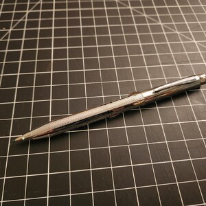 Peut inclure: Un stylo argenté avec un corps texturé à motif de losanges et un dessus lisse et brillant. Le stylo est posé en diagonale sur un tapis de coupe noir avec un motif quadrillé. La pointe du stylo est visible.