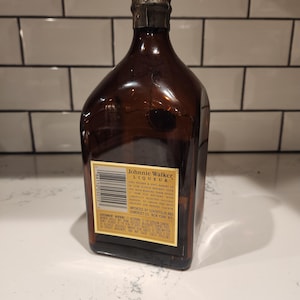 Pode incluir: Uma garrafa de vidro castanho de licor Johnnie Walker. A garrafa tem uma cor castanha escura e uma tampa met&aacute;lica. O r&oacute;tulo inclui o nome da marca e outros textos. A garrafa est&aacute; numa superf&iacute;cie branca.