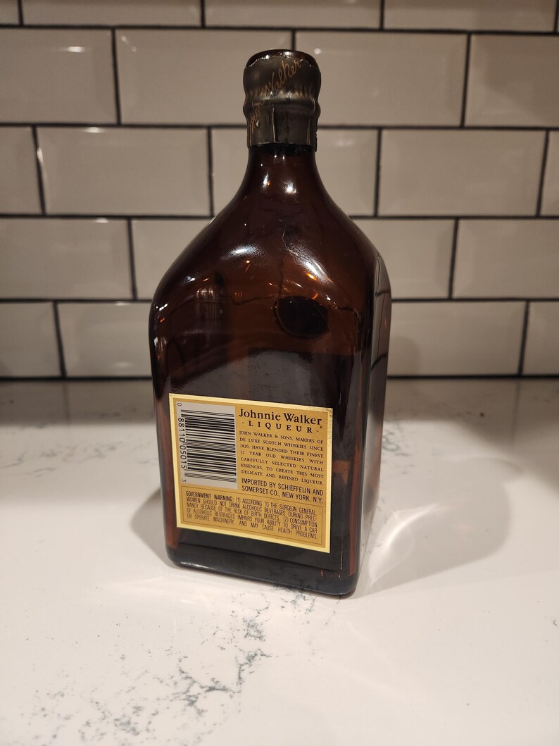 Pode incluir: Uma garrafa de vidro castanho de licor Johnnie Walker. A garrafa tem uma cor castanha escura e uma tampa met&aacute;lica. O r&oacute;tulo inclui o nome da marca e outros textos. A garrafa est&aacute; numa superf&iacute;cie branca.