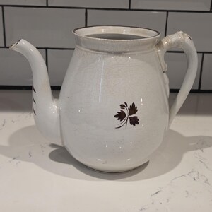 May include: A cream-colored ceramic teapot featuring a subtle crackle glaze finish. The body is adorned with a small, dark brown leaf motif on the front. It includes a curved spout, a sturdy handle, and a wide opening at the top with a thin rim.
