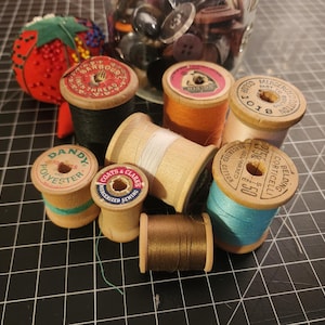 May include: A collection of vintage sewing supplies, including spools of thread in various colors, a red strawberry pincushion, and a jar of buttons. The spools have labels with brand names and thread type. The thread colors include black, orange, teal, and brown.