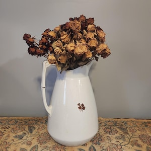 May include: A white ceramic pitcher with a handle, filled with dried brown roses. The pitcher has a brown leaf design on the front. The pitcher is sitting on a floral patterned fabric.