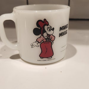 May include: White ceramic mug featuring a Minnie Mouse graphic. Minnie is depicted in a red dress, red bow, and striped shirt. The mug has a handle and the words "MINNIE MOUSE" are printed on the side. The mug is a vintage collectible.