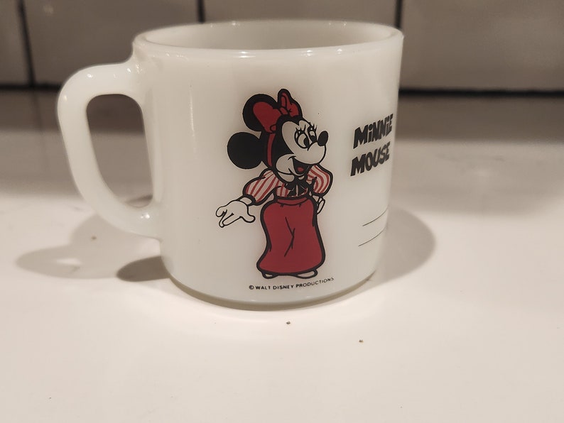 May include: White ceramic mug featuring a Minnie Mouse graphic. Minnie is depicted in a red dress, red bow, and striped shirt. The mug has a handle and the words "MINNIE MOUSE" are printed on the side. The mug is a vintage collectible.