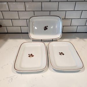 May include: Set of three white rectangular serving dishes with a brown trim and a brown leaf design in the center. The dishes are arranged on a white countertop in front of a white tile backsplash.