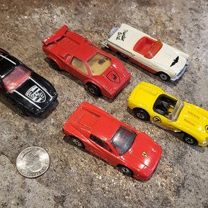 May include: A collection of toy cars, including a black Porsche, red Lamborghini, white and red convertible, red Ferrari, and yellow Ferrari. The cars are arranged on a textured, gray surface, with a quarter coin visible.