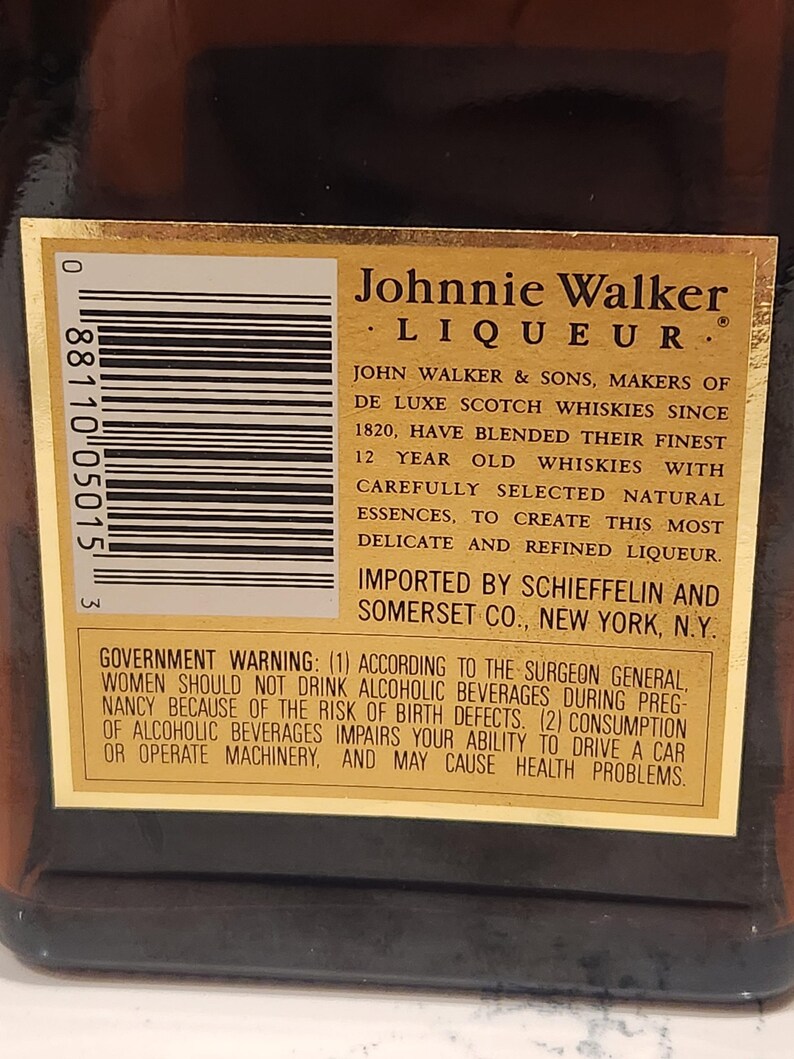 Pode incluir: Close-up de um r&oacute;tulo dourado em uma garrafa marrom de licor Johnnie Walker. O r&oacute;tulo apresenta texto preto detalhando o produto e um aviso governamental. A marca &eacute; proeminente.