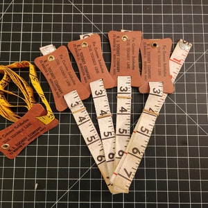 May include: A selection of vintage measuring tapes and tags. The tags are brown and feature the text "R.I. Toomey Company" and "The Toomey Line". The measuring tapes are white with black markings, showing measurements in inches.