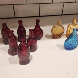 May include: A collection of small, decorative glass bottles in various shapes and colors. The bottles are primarily shades of red, amber, and blue. Some bottles have textured surfaces, while others are smooth. The bottles are arranged on a white surface.