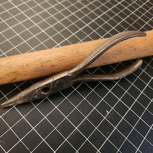 May include: Close-up of a pair of needle-nose pliers with a textured handle, resting on a wooden dowel. The pliers are made of metal and have a pointed tip. The background is a black grid.