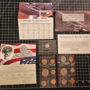 May include: A collection of United States Mint uncirculated coin sets, including a 1996 set, displayed on a black grid surface. The sets include various coins in protective packaging, along with informational cards and certificates.
