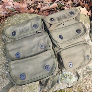 May include: Olive green canvas military pouches with the letters "U.S." embossed on the flaps. Each pouch has a series of pockets and is secured with black snap closures. The pouches are arranged on a stone surface.