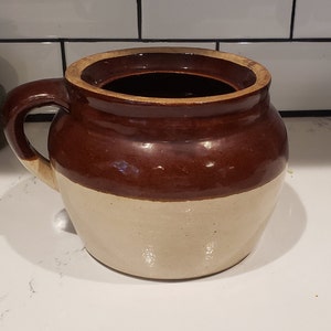 May include: A two-toned ceramic crock with a handle. The upper portion is a rich brown, while the lower half is a creamy off-white. The crock has a wide opening and a sturdy handle, suggesting it's designed for storage or serving.
