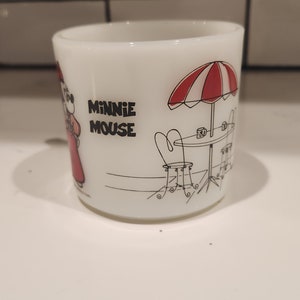 May include: White ceramic mug featuring Minnie Mouse in a red dress. The mug has a black and white illustration of a cafe scene with a red and white striped umbrella and the words "MINNIE MOUSE".