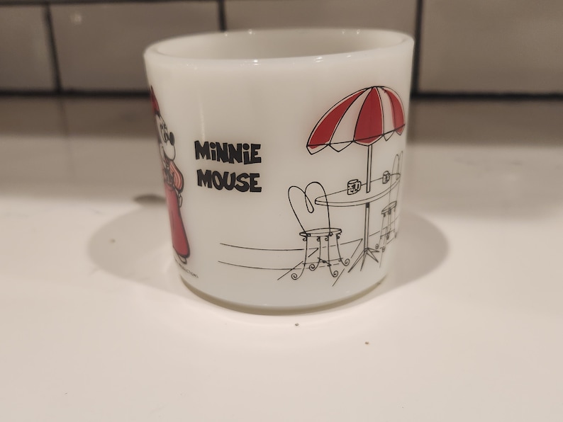May include: White ceramic mug featuring Minnie Mouse in a red dress. The mug has a black and white illustration of a cafe scene with a red and white striped umbrella and the words "MINNIE MOUSE".