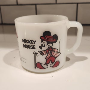 May include: White ceramic mug featuring Mickey Mouse in a red and white striped suit, hat, and cane. The mug has a handle and the words "Mickey Mouse" printed on the side. A vintage collectible.