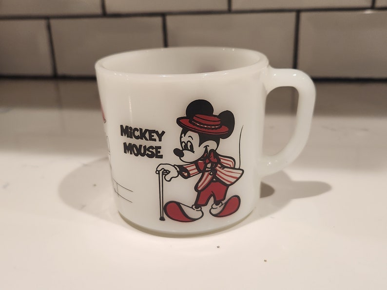 May include: White ceramic mug featuring Mickey Mouse in a red and white striped suit, hat, and cane. The mug has a handle and the words "Mickey Mouse" printed on the side. A vintage collectible.