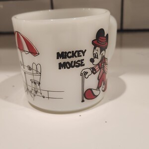 May include: White ceramic Mickey Mouse mug. The mug features a black and red illustration of Mickey Mouse in a red suit and hat, with a cane. The mug also has the words "Mickey Mouse" in black text.