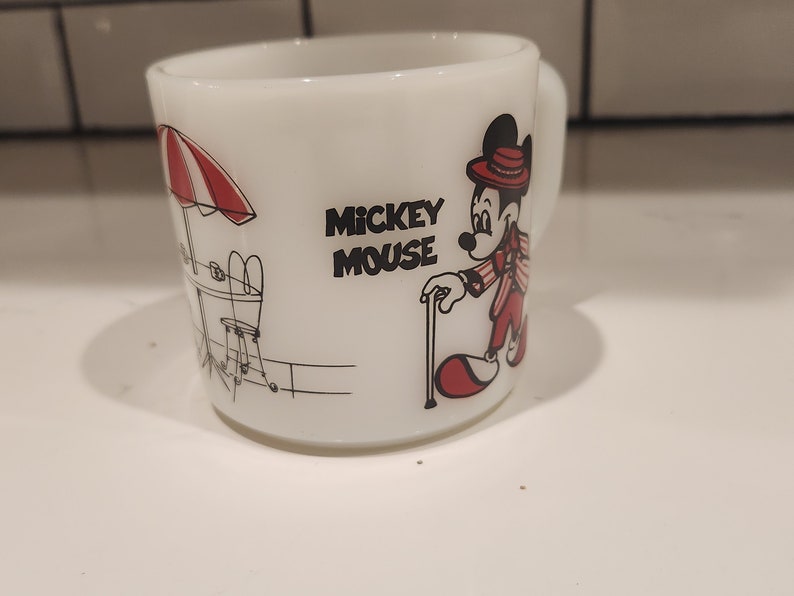 May include: White ceramic Mickey Mouse mug. The mug features a black and red illustration of Mickey Mouse in a red suit and hat, with a cane. The mug also has the words "Mickey Mouse" in black text.
