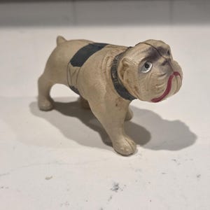 May include: A cast iron bulldog figurine painted in a light beige color with a black collar and a black patch on its back. The dog has detailed facial wrinkles, dark shading around the eyes, and a red painted mouth, standing in a fixed pose.