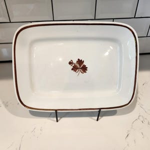 May include: A rectangular white serving dish with a brown border and a brown leaf design in the center. The dish is displayed on a black metal stand, suitable for serving food at a gathering.