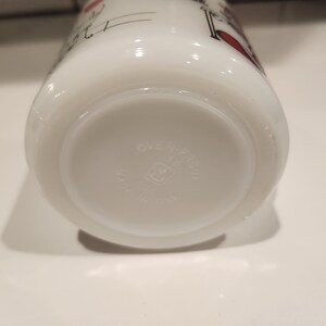 May include: White Pyrex mug with a design featuring black and red accents. The bottom of the mug has the text "OVEN-PROOF" and "PYREX" in a circular pattern. The mug is made in the USA.