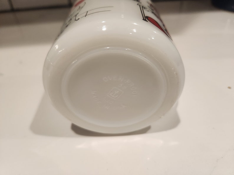May include: White Pyrex mug with a design featuring black and red accents. The bottom of the mug has the text "OVEN-PROOF" and "PYREX" in a circular pattern. The mug is made in the USA.