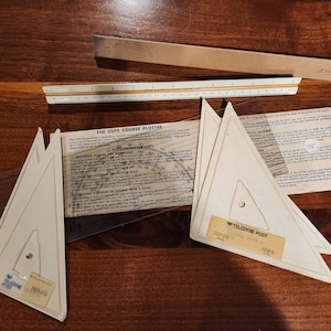 May include: A selection of vintage drafting instruments, including a wooden ruler, a scale, and several triangular course plotters. The plotters are labelled "Teledyne Post" and the text "The USPS Course Plotter" is visible on a paper. The items are arranged on a wooden surface.