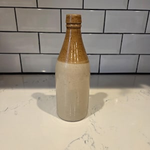 May include: A vintage stoneware bottle with a two-tone design. The top portion is a warm brown, transitioning to a light beige base. The bottle has a narrow neck and a rounded body, suggesting it may have been used for storing liquids.