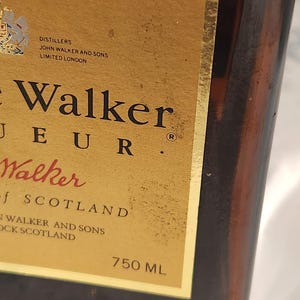 Pode incluir: Close-up de uma garrafa de vidro castanho com um r&oacute;tulo dourado. O r&oacute;tulo apresenta as palavras "John Walker" e "750 ML". A garrafa &eacute; parcialmente vis&iacute;vel, mostrando sua cor &acirc;mbar escuro. O r&oacute;tulo tamb&eacute;m inclui o texto "Distillers, John Walker and Sons, Limited London."