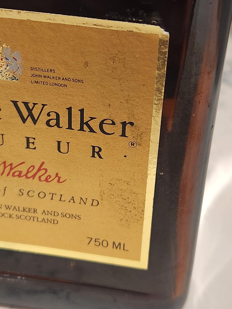 Pode incluir: Close-up de uma garrafa de vidro castanho com um r&oacute;tulo dourado. O r&oacute;tulo apresenta as palavras "John Walker" e "750 ML". A garrafa &eacute; parcialmente vis&iacute;vel, mostrando sua cor &acirc;mbar escuro. O r&oacute;tulo tamb&eacute;m inclui o texto "Distillers, John Walker and Sons, Limited London."