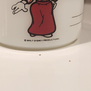 May include: White ceramic mug featuring a cartoon character in a red skirt and a white glove. The mug has a handle and the text "WALT DISNEY PRODUCTIONS" printed on the bottom.