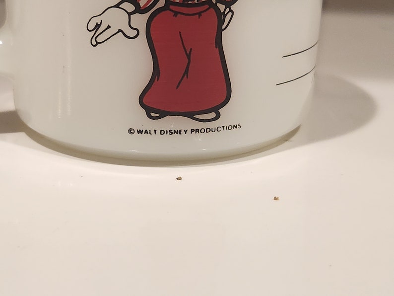 May include: White ceramic mug featuring a cartoon character in a red skirt and a white glove. The mug has a handle and the text "WALT DISNEY PRODUCTIONS" printed on the bottom.